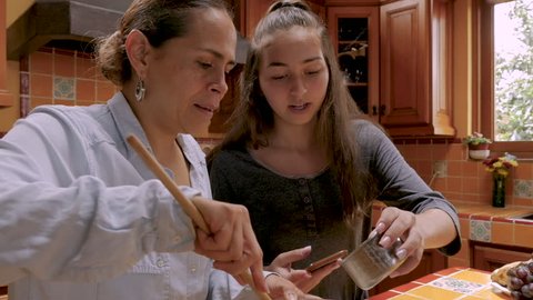 Happy smiling teenager girl referencing an app on her cell phone to help her Mexican mother bake in their kitchen together - slow motion push in