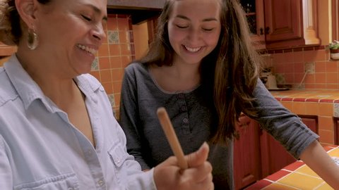 Hispanic mother and teenager daughter look at each other smiling while cooking together - slow motion