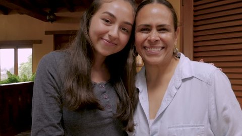 Happy Mexican teenager daughter kissing her mother on the cheek and then smiling and looking at the camera in their home - slow motion