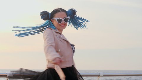 Portrait of beautiful girl with african kanekalon blue braids and heart shaped sunglasses dancing on sea background. Afro hairstyle, freaky dyed hair. Hipster having fun.