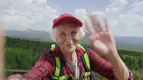 Happy senior woman with hiking backpack smiling and waving at camera while talking via video call from mountain viewpoint