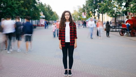 Zoom in time-lapse of beautiful girl with long curly hair standing in street looking at camera when many men and women are walking around in hurry on summer day.