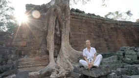 Young woman exercising yoga in ancient temple near majestic tree roots; sunlight passing through and relaxing with some meditation; concept of people with healthy lifestyles wellbeing travel 4K
