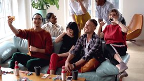 Joyful trendy dressed male and female hipsters taking image of common rest in apartment sitting on sofa, smiling guy using mobile phone for making selfie with friends recreating on party in flat