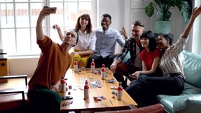 Smiling trendy dressed man making funny selfie with his friends on smartphone resting on home party together, cheerful group of multiracial hipsters posing for image while playing cards and recreating