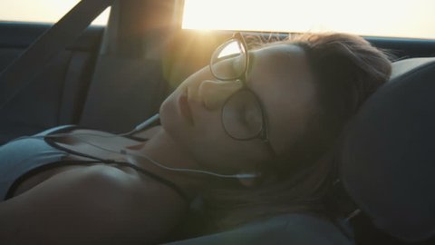 A girl with glasses rides in the car in the front seat and sleeps with headphones in her ears. Slow motion.