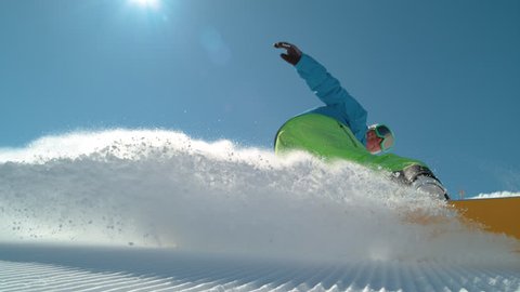 SLOW MOTION CLOSE UP: Extreme snowboarder carving downhill and spraying snow into camera. Smiling active man snowboarding on groomed ski slope in mountain resort. Cheerful snowboarder turning