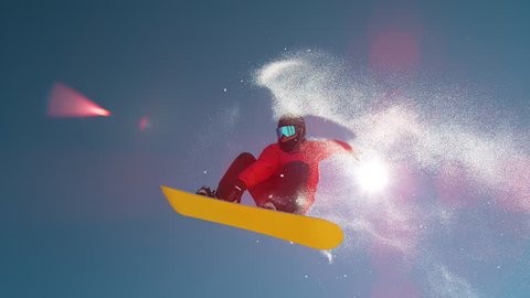 SLOW MOTION CLOSE UP: Snowboarder jumping big air kicker, spraying snowflakes and flying over sun on perfect winter day. Snowboard jump in snow park. Sunbeams shining past jumping boarder in mountains