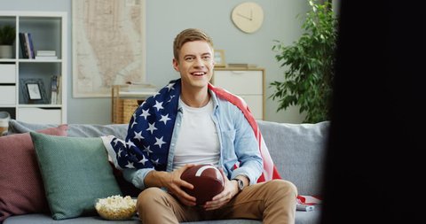 Caucasian young man with American flag and rugby ball watching sport game on TV and being sad of loosing. 库存视频