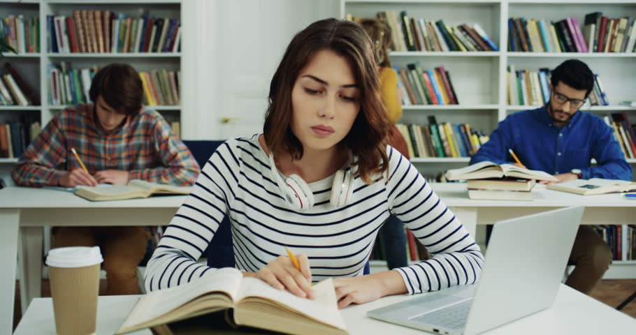 Pretty young woman with headphones studing in the library with books, coffee and laptop, then smiling and lokking to the camera.