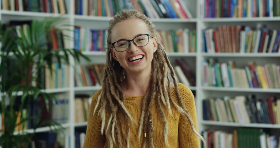 Portrait shot of the young hipster woman in glasses and with dreadlocks laughing in front of the camera in the library.