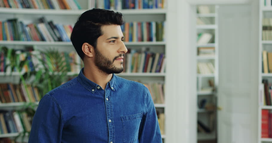 Portrait shot of the Arabian handsome young man standing in the modern library, looking at the side and turning head to the camera with a smile.