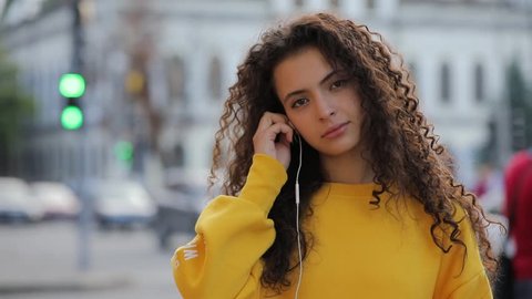 Portrait of young multiracial girl in yellow pullover puts on earphones in urban city background