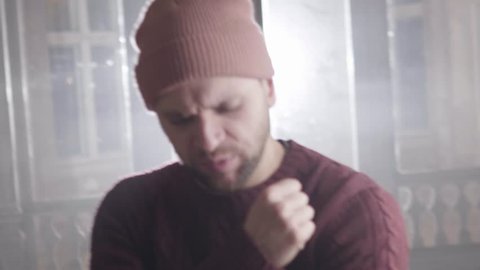 Young bearded man dressed in brown knitted sweater and pastel sock cap is rapping with hand gesturing, looking at camera and stepping back and forward during rap in bright room.