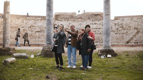 Smiling senior family and young woman tourists waving on video call to family at old amphitheater ruins in Ostia, Italy.