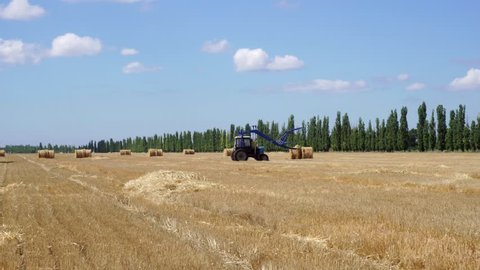 Modern technology agricultural tractor collecting huge bales of hay from big organic wheat field on summer day landscape