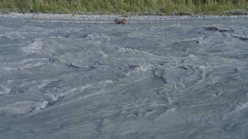 Slow-motion pan up from dirty water flowing in the Matanuska River outside of Anchorage, Alaska, to green forest below mountain peak.