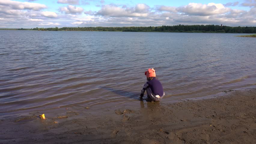 A little girl 4-5 years old plays in the mud on the river bank. A child with blond hair. She wet her hands and clothes in cold water.