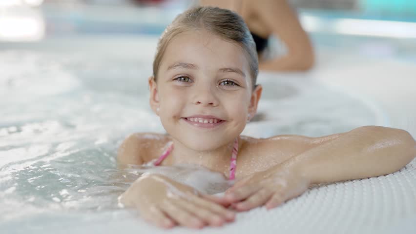 pretty little girl with wet hair is sitting alone in hydromassage bath in a water park, looking to camera