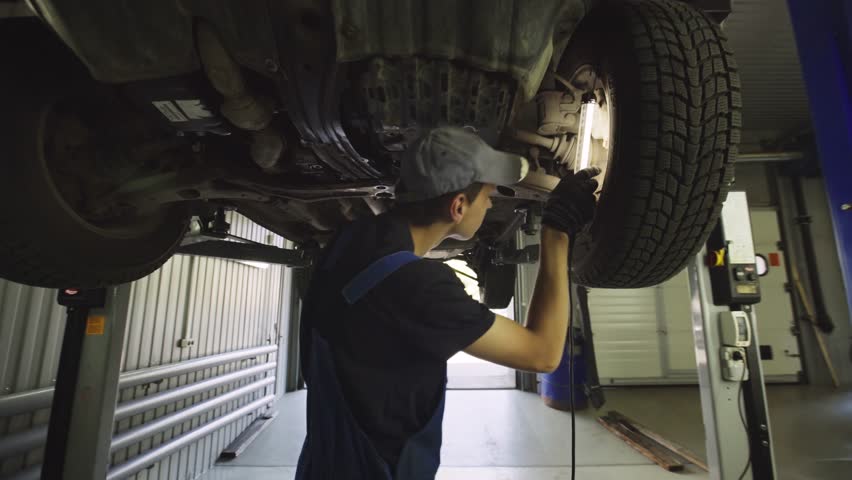 Young mechanic with light in hands looking at steering chassis under a car. Garage service. Automobile diagnostic. Auto repair concept | Shutterstock HD Video #1016433370