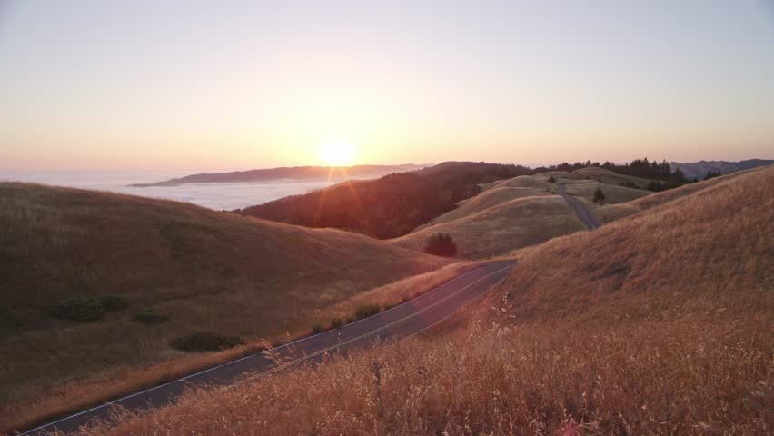 A white car drives down an isolated road, while the sun sets over a beautiful grassy meadow along the California coast