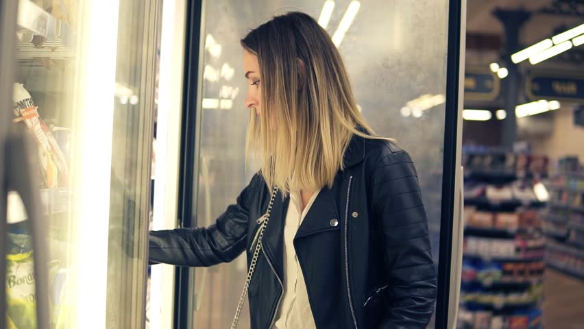 Attractive woman choosing the dairy products in the supermarket in refrigerator. Young woman takes frozen products, vegetablels to a shop basket. Close up