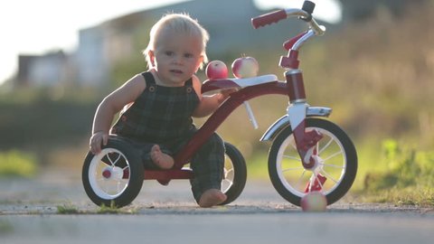 Cute toddler child, boy, playing with tricycle in park and eating apple, kid riding bike on sunset
