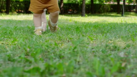 Sideways tracking with low-section of unrecognizable mother teaching baby to walk on green grass in park on summer day