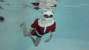 A little boy dressed as Santa Claus goes in for sports with a coach in the pool. He swims underwater to the stairs in swimming goggles and looks into the camera. Slow motion. Shooting underwater.