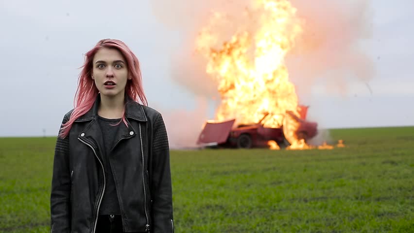 Young beautiful girl with pink hair and in a leather jacket stands on the background of an exploding car, a hipster and a burning car, car explosion