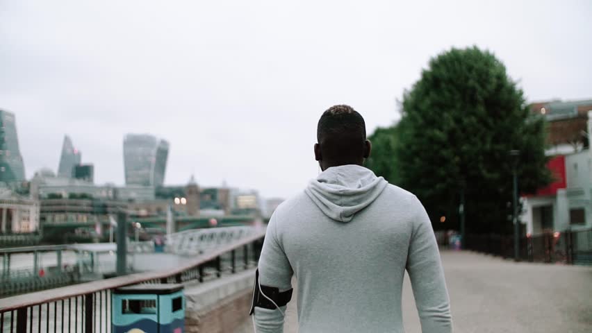 A rear view of young sporty black man runner with smart phone walking on the bridge in a city.