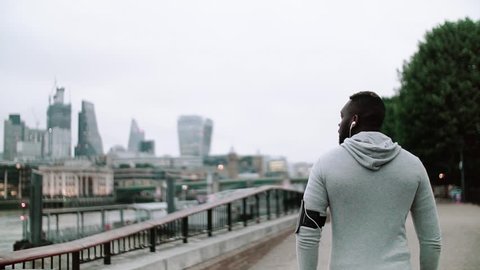 A rear view of young sporty black man runner with smart phone walking on the bridge in a city.