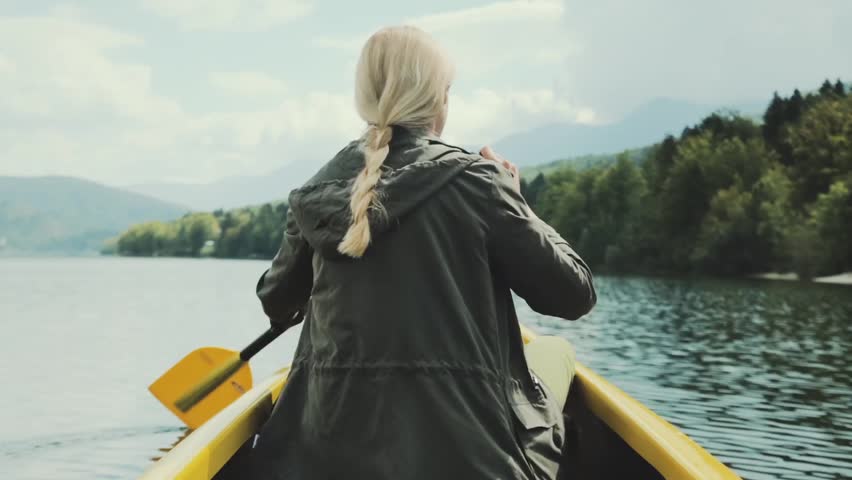 Girl traveler swims in boat on mountain lake surrounded by high mountains. Girl in canoe rowing leisurely paddle along calm lake. Woman in the boat from the back.