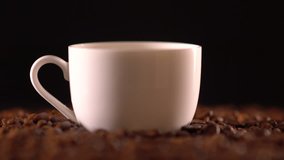 Coffee pouring from metal container into small white mug sitting on mound of whole coffee beans in front of black background