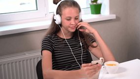 Young girl waving and smiling at her mobile phone as she sits at a table indoors listening to music on stereo headphones