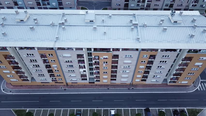 building, windows and terraces, parking, street and car. The car passing by empty street, next to the building and parking space. New building with lots of windows and balconies.