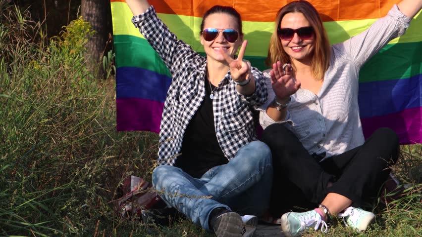 Two young women are sitting on a background of the rainbow flag. The sun is shining brightly, LGBT rights, lesbian family.