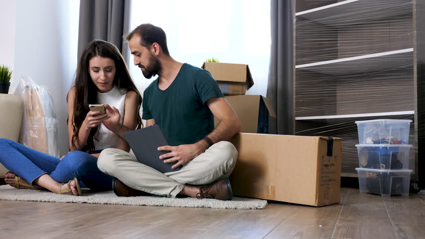 Beautiful young couple sitting on the floor of their new apartment shopping online for new furniture. They are surrounded by cardboard boxes