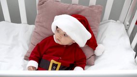 surprised adorable baby in santa costume looking away and sitting in crib at home, christmas concept