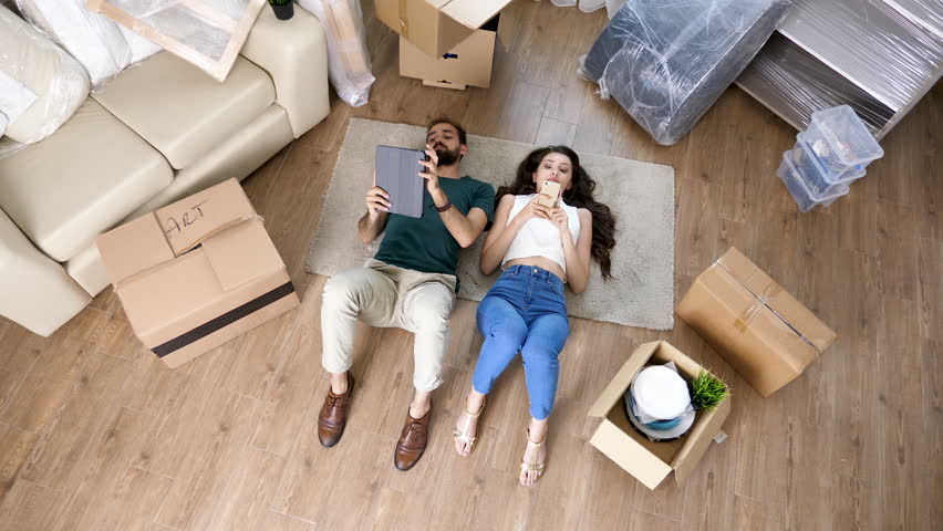 Top view couple lying on the floor of new apartment next to cardboard boxes. They just moved in in their new home
