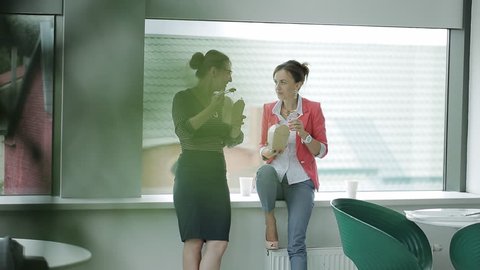 Two business women on a lunch break at the window in the cafeteria eating Chinese food and drinking coffee. The end of the lunch break, the women hurry to take their jobs.