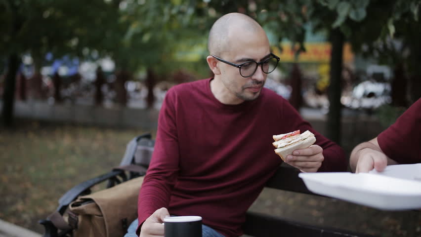Two men having lunch on a Park bench with a sandwich and drink from cups