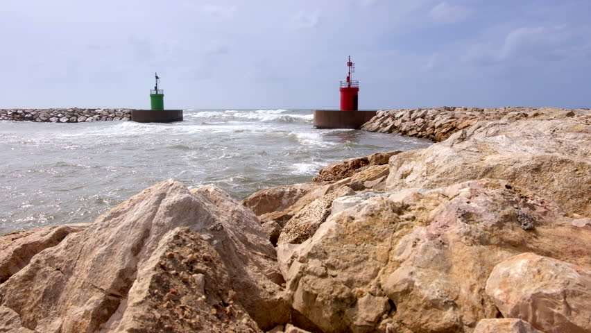 Sea water current and waves on a harbor access with green lighthouse tower viewed from the rocks of a wave safety barrier.