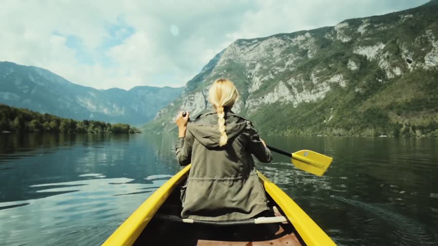Girl traveler swims in boat on mountain lake surrounded by high mountains. Girl in canoe rowing leisurely paddle along calm lake. Woman in the boat from the back. Royalty-Free Stock Footage #1016600233