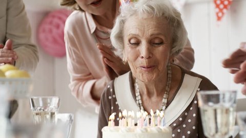 Beautiful senior woman  in party hat blowing off birthday candles on cake while joyous friends clapping hands and saying best wishes