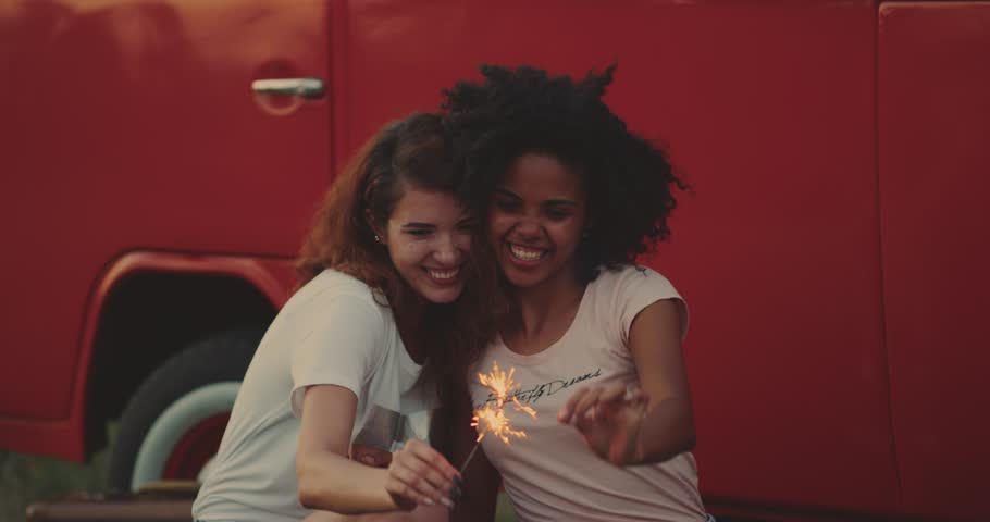 One african young female and her friend , bought with curly hair smiling playing with a sparklers at the picnic sitting down on the grass beside of a retro van. 4k.
