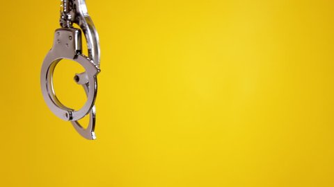 Shaking a pair of shiny metal handcuffs over a yellow background, room for copy-space on the right. Symbolic shot: crime, punishment, menace, fetish, sexuality, pleasure.
