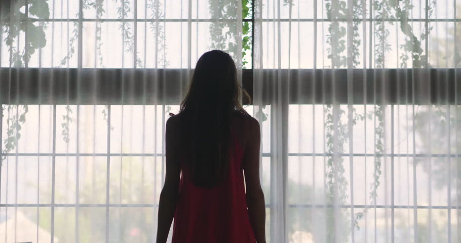 Portrait of a young woman in a red dress   is opening a curtain the let the light come through the window. Royalty-Free Stock Footage #1016623882
