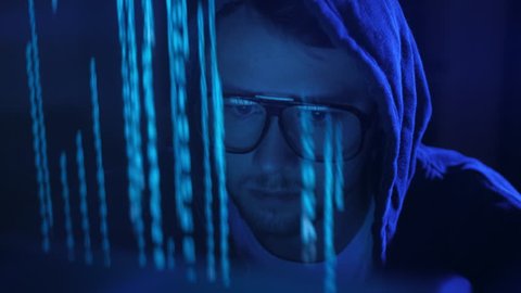Close up of generation z hacker in glasses and hoodie working on computer under the blue neon light sparkle. Darkness background. Cybersecurity concept. ProRes 422