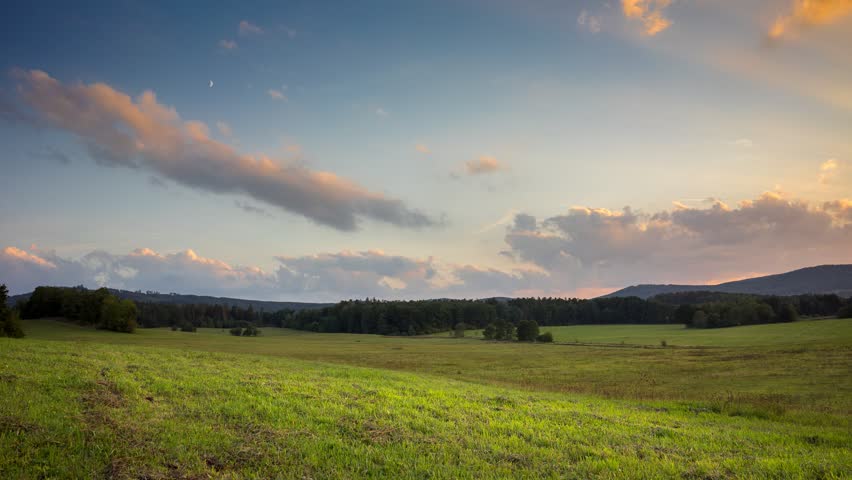 Green meadows and small hills under the gold and blue sunset with clouds. Timelapse video.